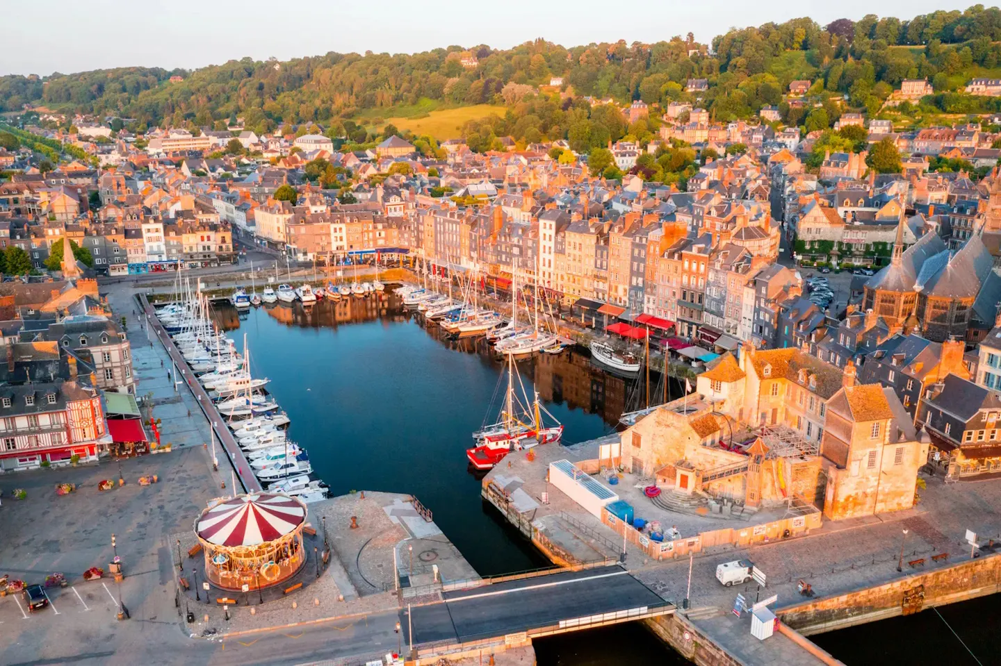 Aerial view of Honfleur, France, featuring a harbor filled with sailboats, colorful buildings, and a carousel.