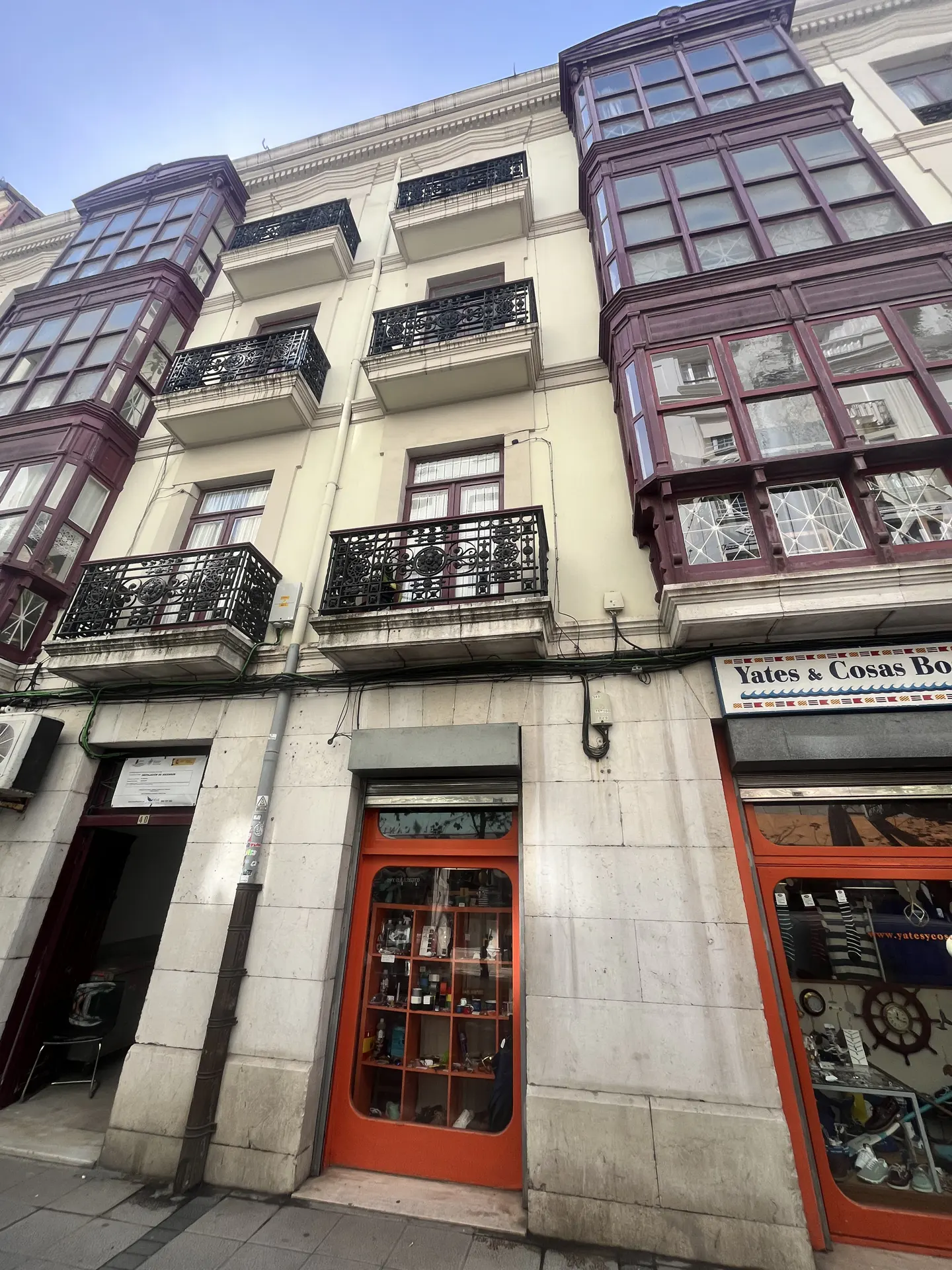Exterior view of a multi-story building with balconies and bay windows above storefronts. The building is cream-colored with red and black accents.