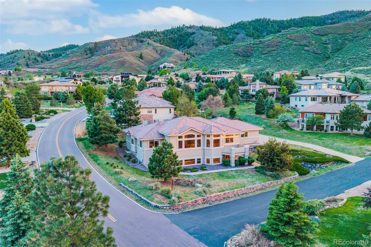 Aerial view of a beige house with a red tile roof, surrounded by green trees and a mountain backdrop.