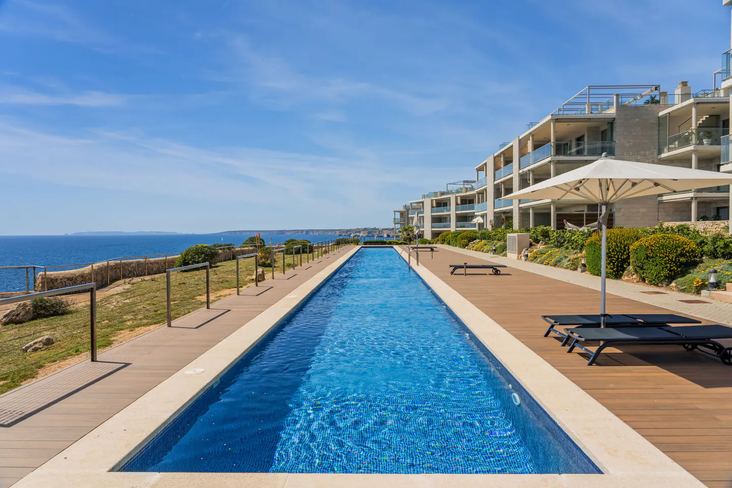 A long, narrow blue pool stretches toward the ocean, lined by condos, lounge chairs, and a white umbrella.