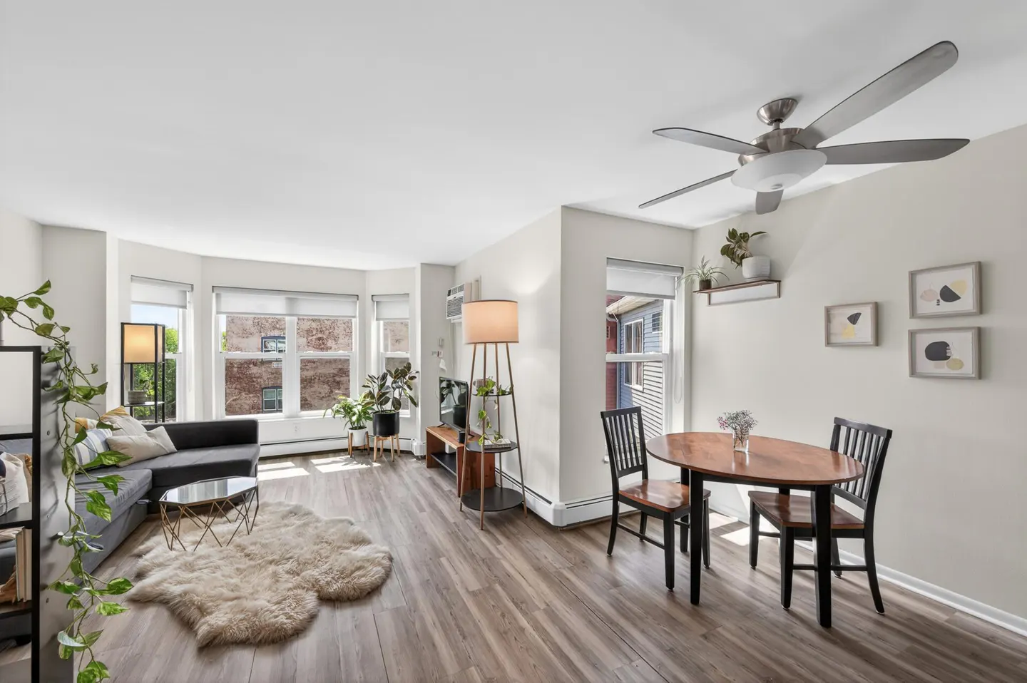 Bright, open apartment living space with gray sofa, wood floors, dining table, and many plants.