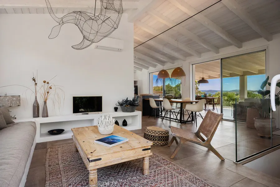Bright, open living space with white walls, a wooden coffee table, and a woven chair. A wire sculpture hangs from the ceiling. Sliding glass doors lead to an outdoor view.