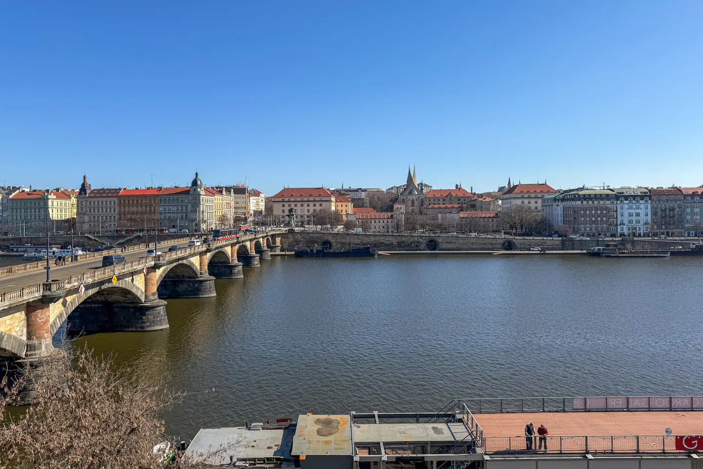 Cityscape view of Prague with a stone bridge over the Vltava River under a clear blue sky. Buildings line the riverbank.