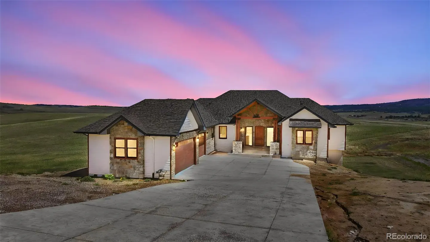 A ranch-style home with stone accents and a gray roof sits on a hill under a pink and purple sunset.