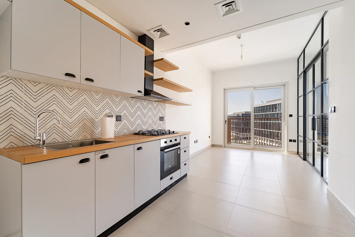 Bright, modern kitchen with gray cabinets, wood countertop, and chevron backsplash. Oven, sink, and open shelves visible. Balcony doors open to city view.