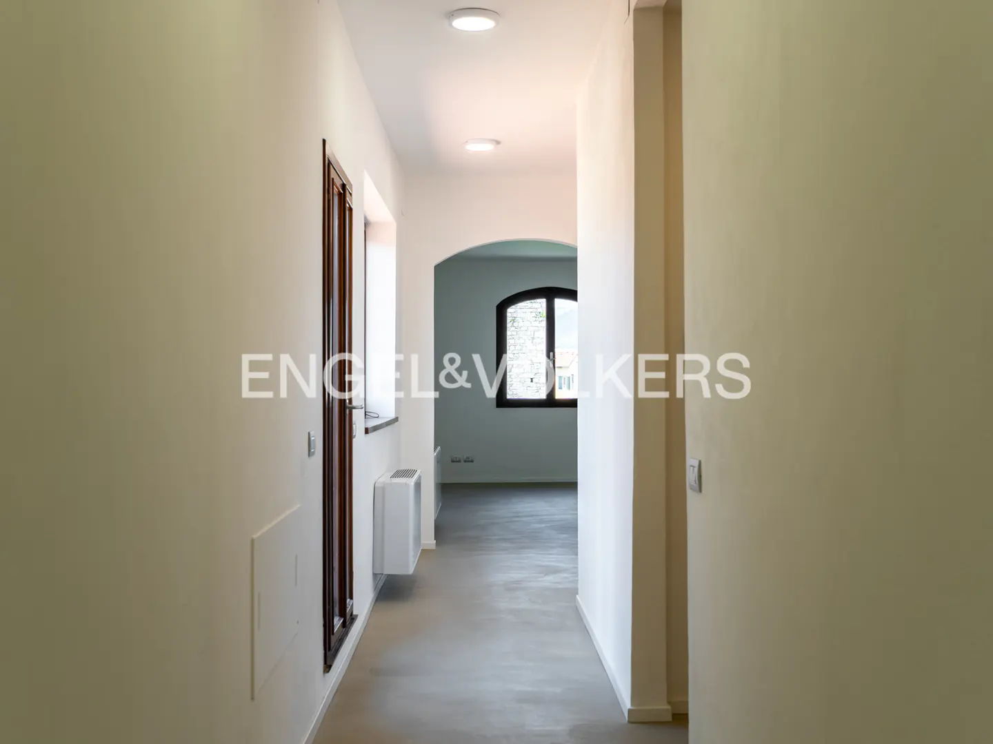 A bright hallway with white walls and a gray floor leads to a room with an arched window. A radiator is under a window.