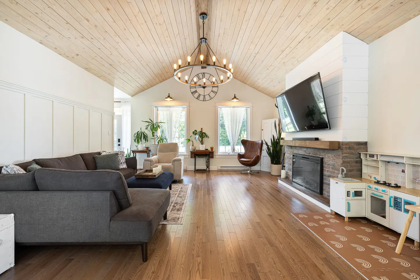Bright living room with wood floors, vaulted ceiling, and a large chandelier. A gray sectional sofa faces a fireplace with a TV above it. A toy kitchen sits nearby.