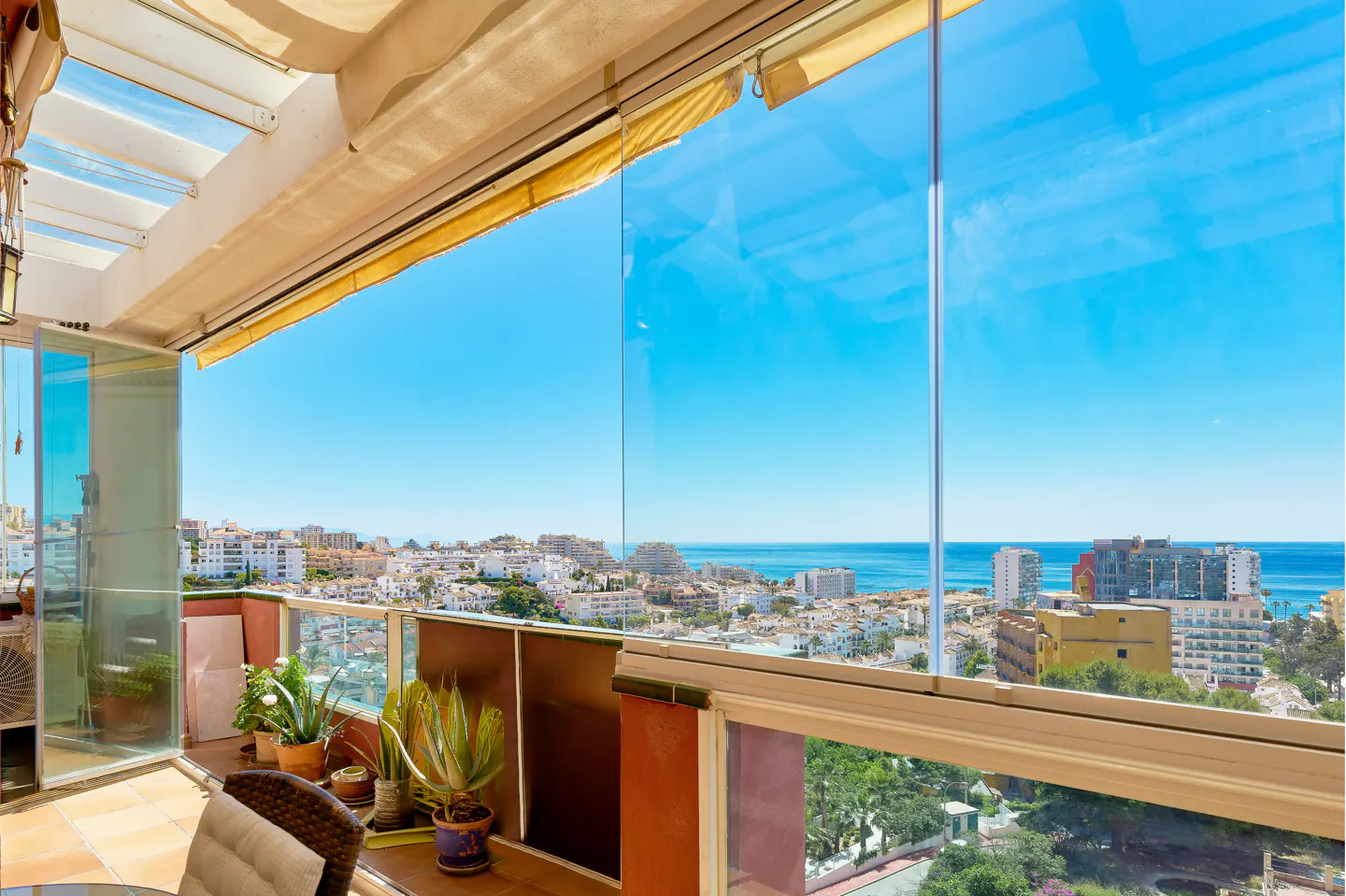 Balcony view with glass walls overlooking a coastal city and the ocean under a clear blue sky. Plants in pots line the balcony.