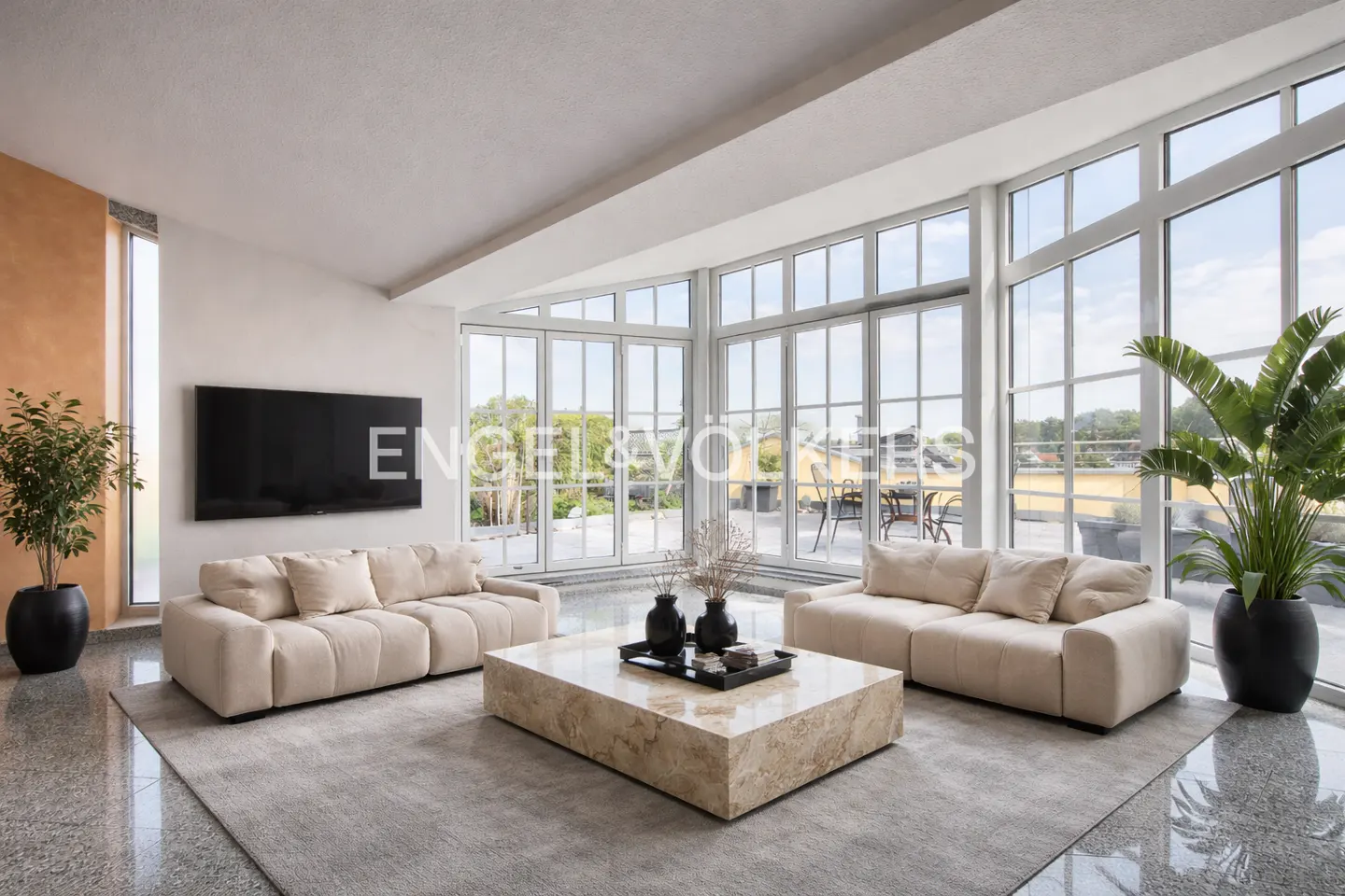Living room with beige sofas, marble table, and floor-to-ceiling windows overlooking a terrace. A large TV is mounted on the wall.