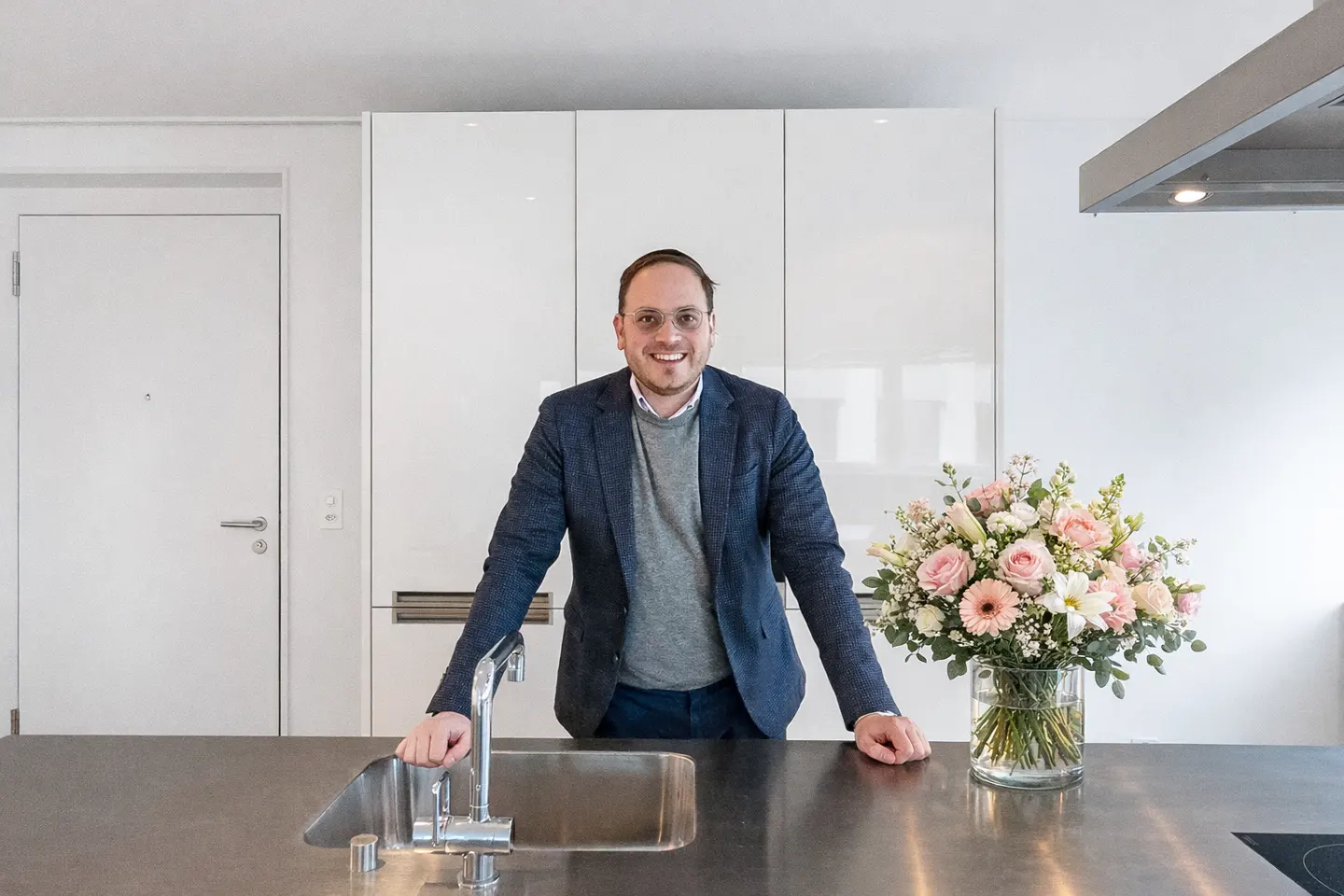 A smiling man in a blazer stands in a modern kitchen with white cabinets and a bouquet of pink and white flowers.
