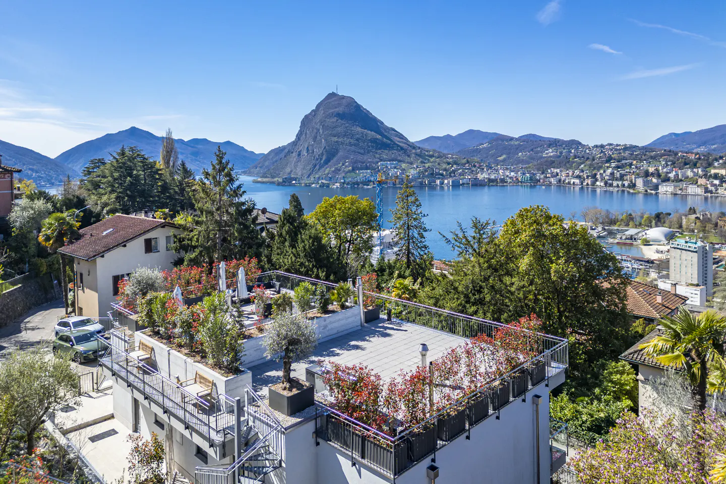 Scenic view of a building with a rooftop garden overlooking Lake Lugano, Switzerland, with mountains in the background under a clear blue sky.