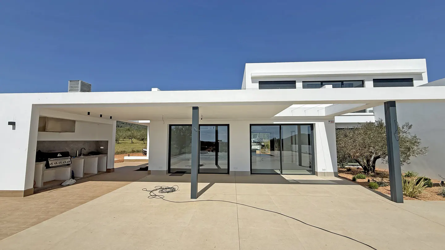 Modern white house exterior with a patio, outdoor kitchen, and sliding glass doors under a clear blue sky.