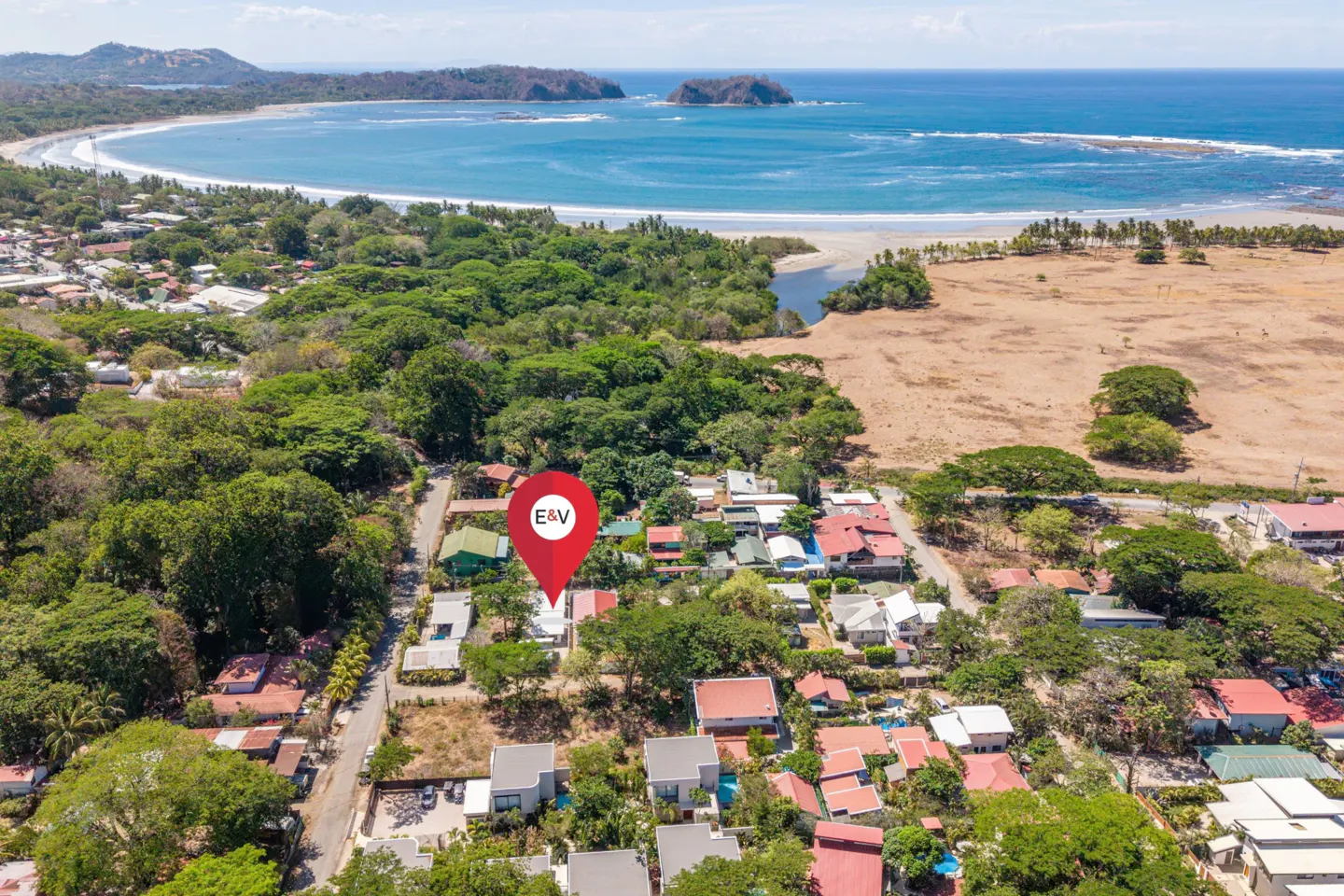 Aerial view of a coastal town with a red location pin over a house, lush green trees, and a sandy beach with blue ocean in the background.