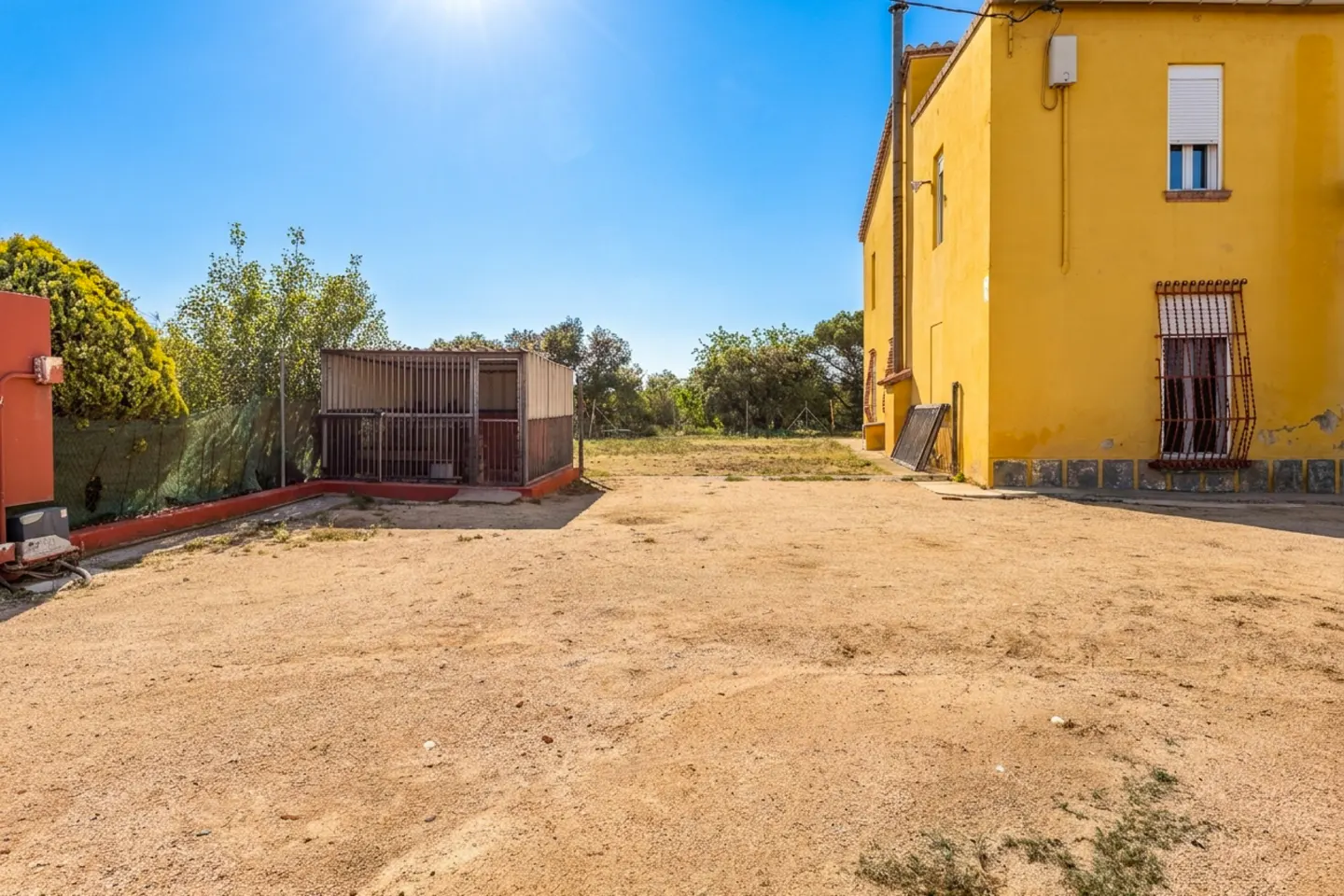 A sunny view of a yellow building with a dirt yard, a metal cage, and trees in the background.