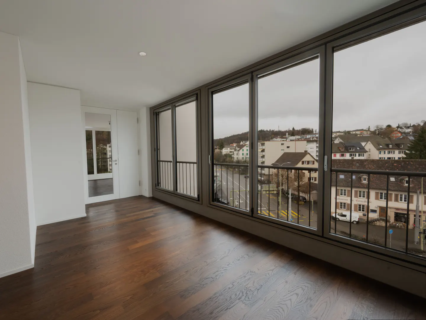 Bright, empty room with dark wood floors and large windows overlooking a town on a cloudy day.