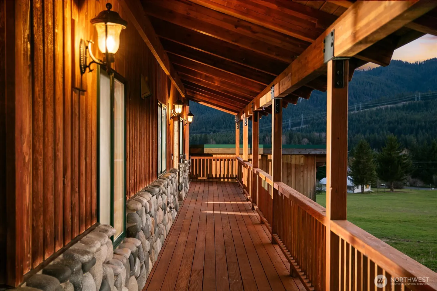 Long wooden porch with stone base and mountain view. Outdoor lights illuminate the wood siding.