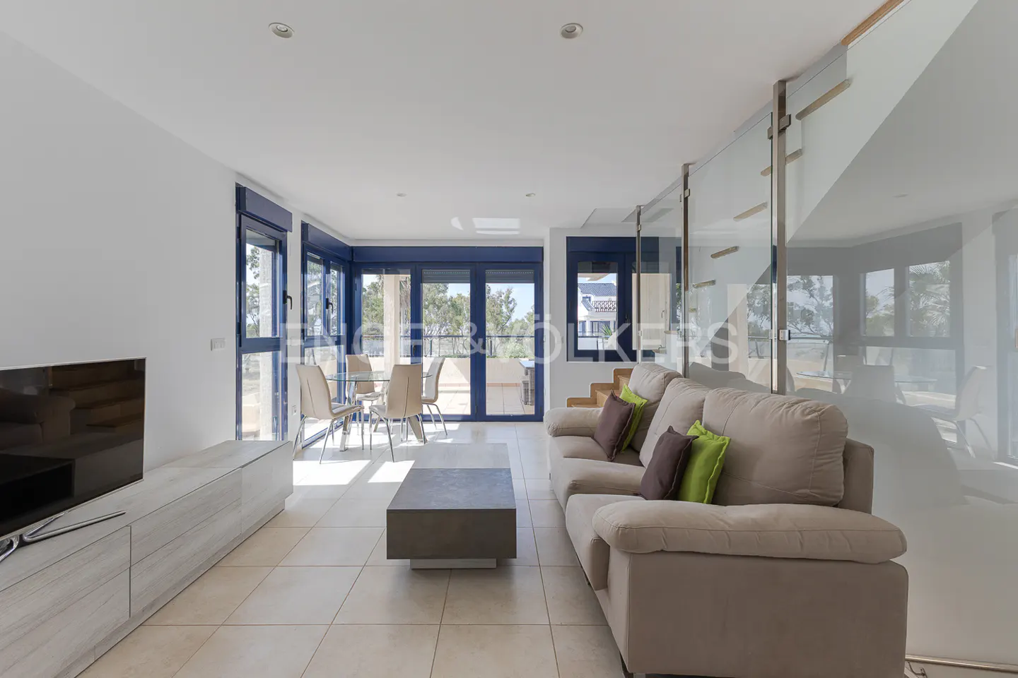 Bright, modern living room with beige sofa, brown and green pillows, gray coffee table, and dining area with blue-framed windows.