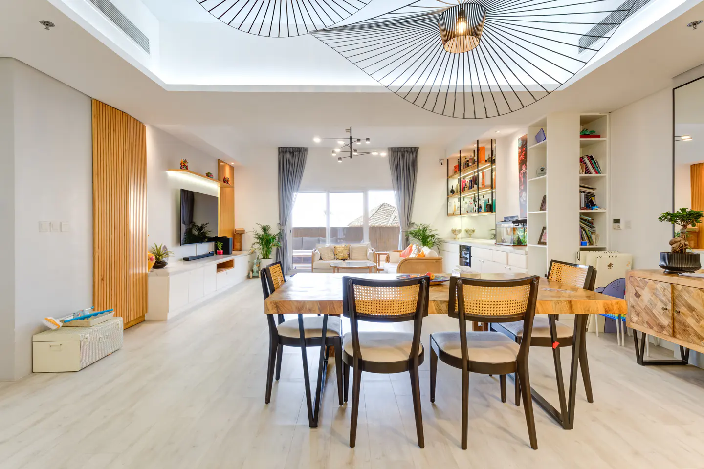 Bright, modern living room with a wood dining table and black chairs. Large windows, white walls, and unique ceiling light fixture.