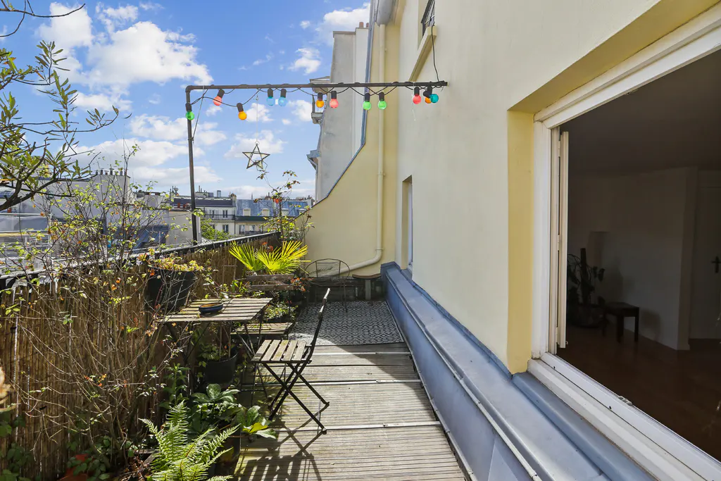 A sunny balcony with a table, chairs, and plants overlooks a city skyline. String lights hang above, adding a festive touch.