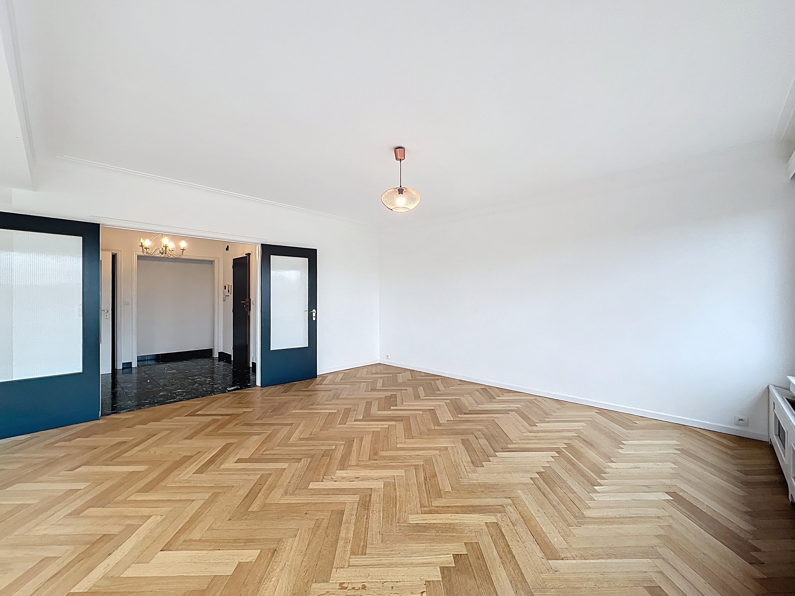 Empty room with herringbone wood floors, white walls, and a glass pendant light. Dark blue sliding doors lead to another room with a chandelier.