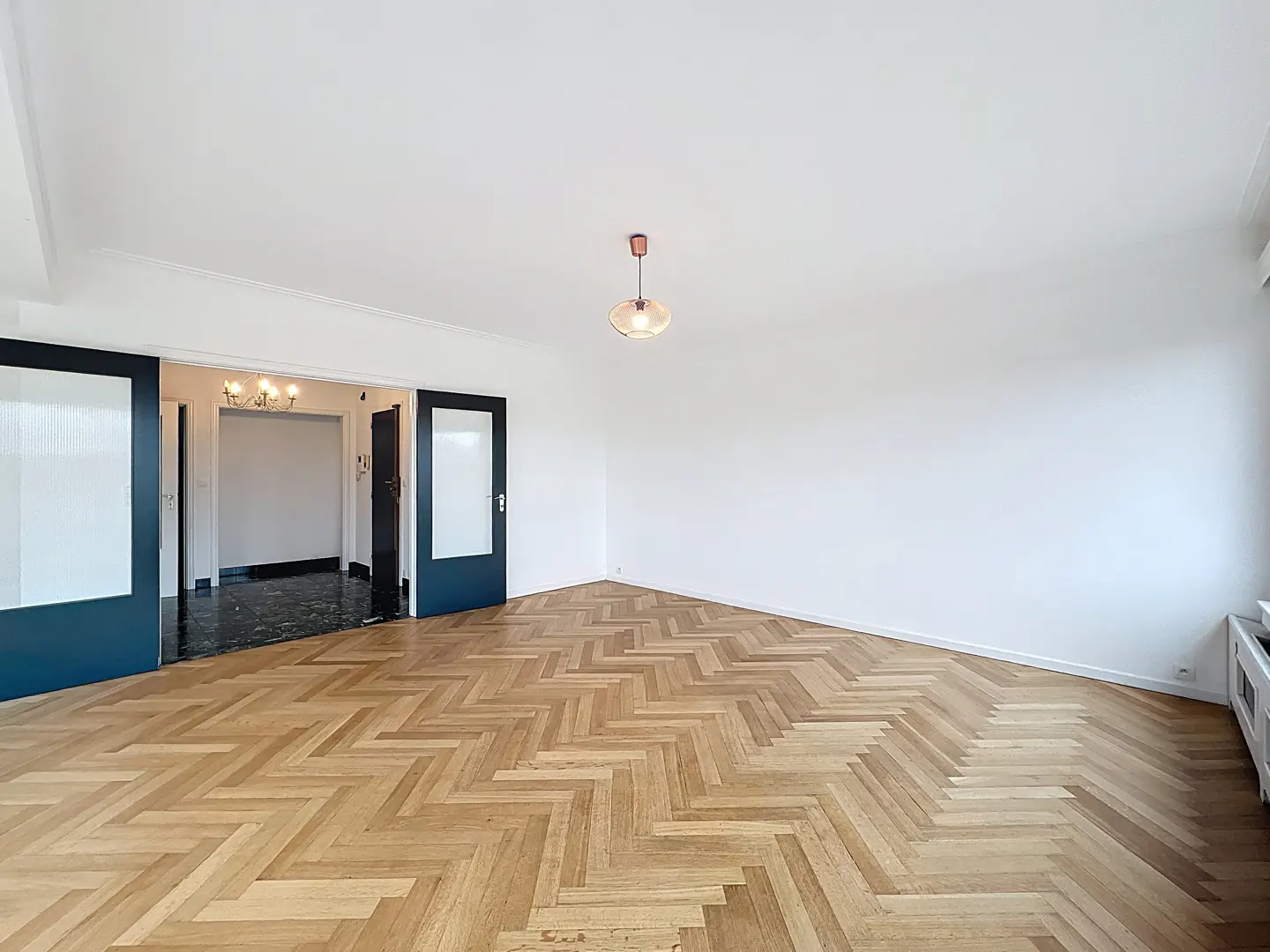 Empty room with herringbone wood floors, white walls, and a glass pendant light. Dark blue sliding doors lead to another room with a chandelier.
