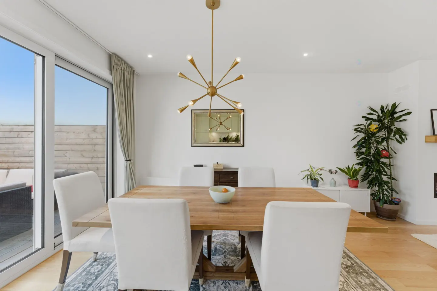 Bright dining room with a wood table, white chairs, and a gold chandelier. Sliding glass doors lead to an outdoor patio.