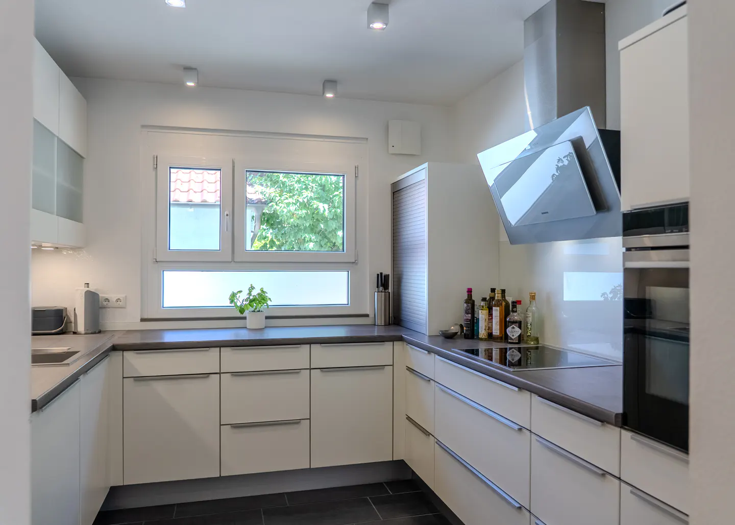 Modern U-shaped kitchen with white cabinets, dark countertops, stainless steel appliances, and a window with garden view.
