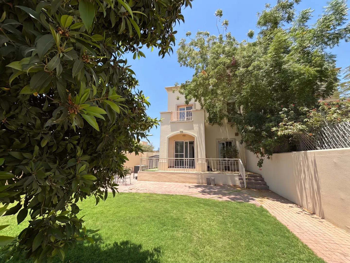 Two-story beige house with a green lawn and trees under a blue sky. A patio with chairs is visible.