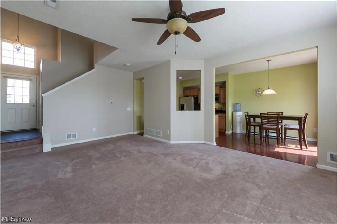 A living room with brown carpet, beige walls, and a ceiling fan. A dining area with a table and chairs is visible through an opening. A staircase leads to the second floor.
