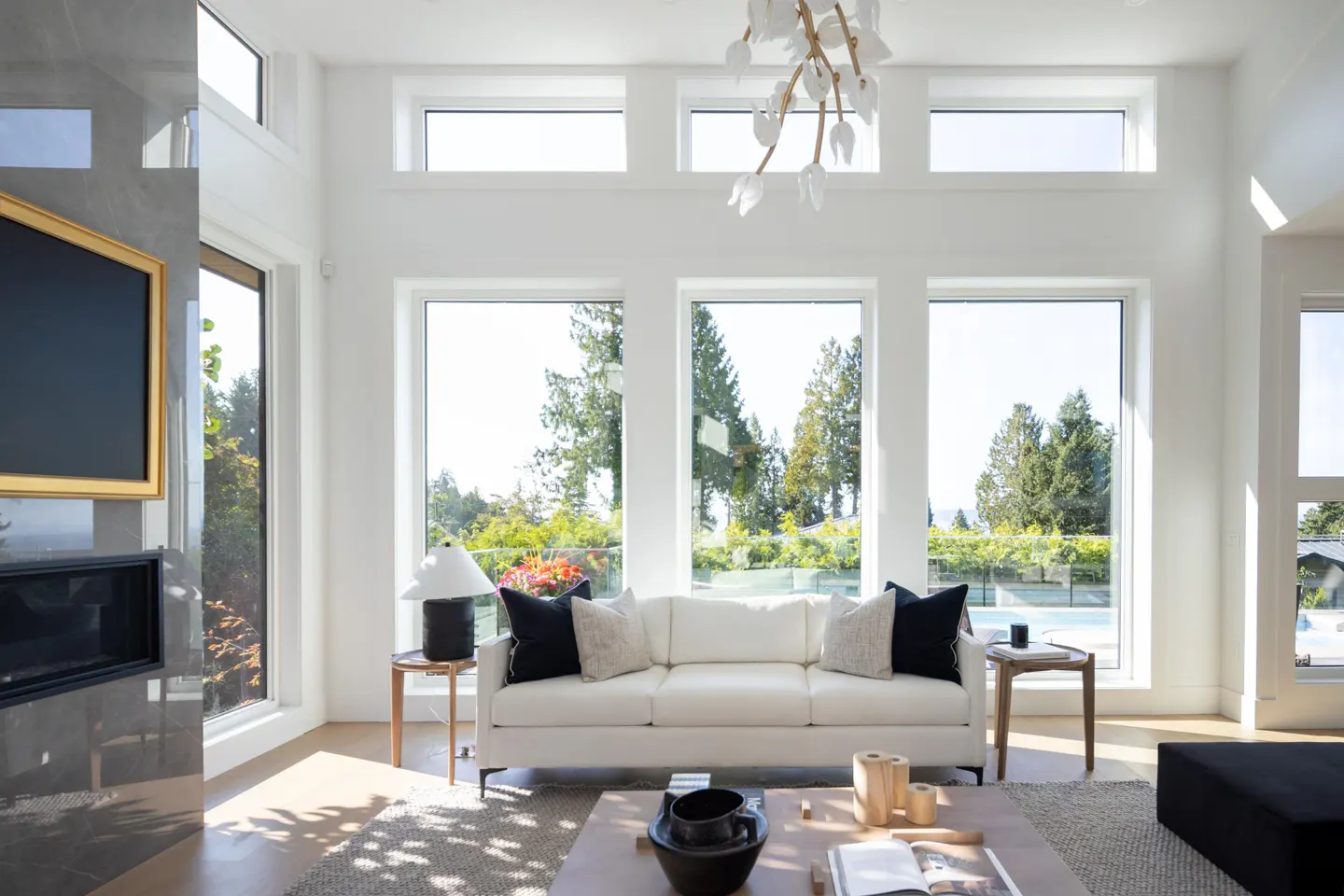 Bright living room with white walls, large windows overlooking trees, and a white sofa with black pillows. A modern chandelier hangs from the ceiling.