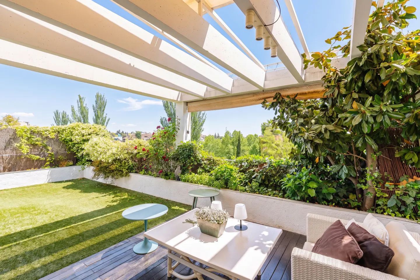 Outdoor patio with white pergola, green lawn, and lush greenery. A white table and wicker sofa with pillows create a cozy seating area.
