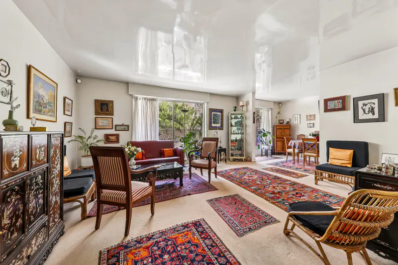 A bright living room with eclectic decor, featuring rugs, a red sofa, and various chairs. Artwork adorns the walls.