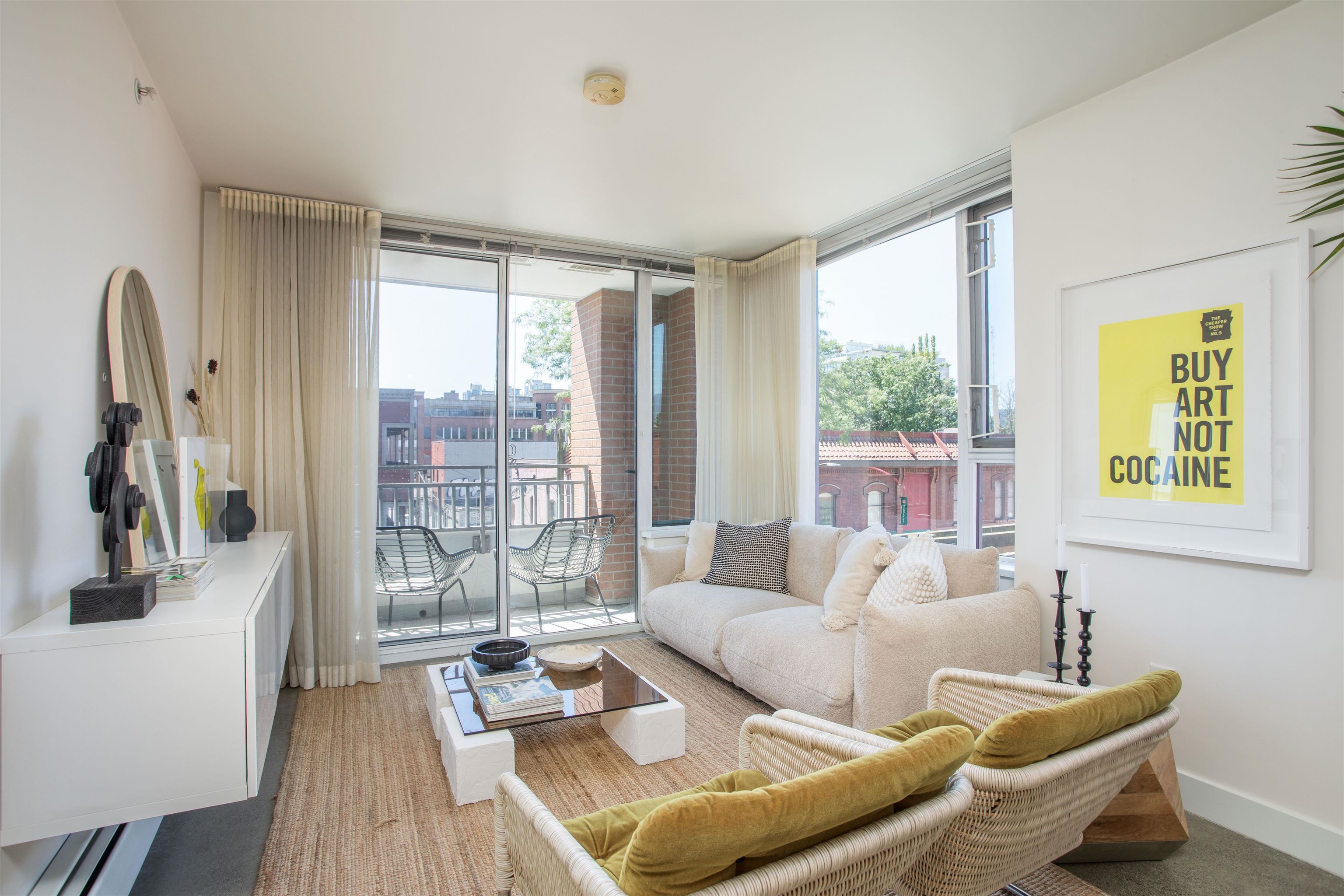 Bright living room with a beige sofa, olive chairs, and a balcony with metal chairs. A "Buy Art Not Cocaine" print hangs on the wall.