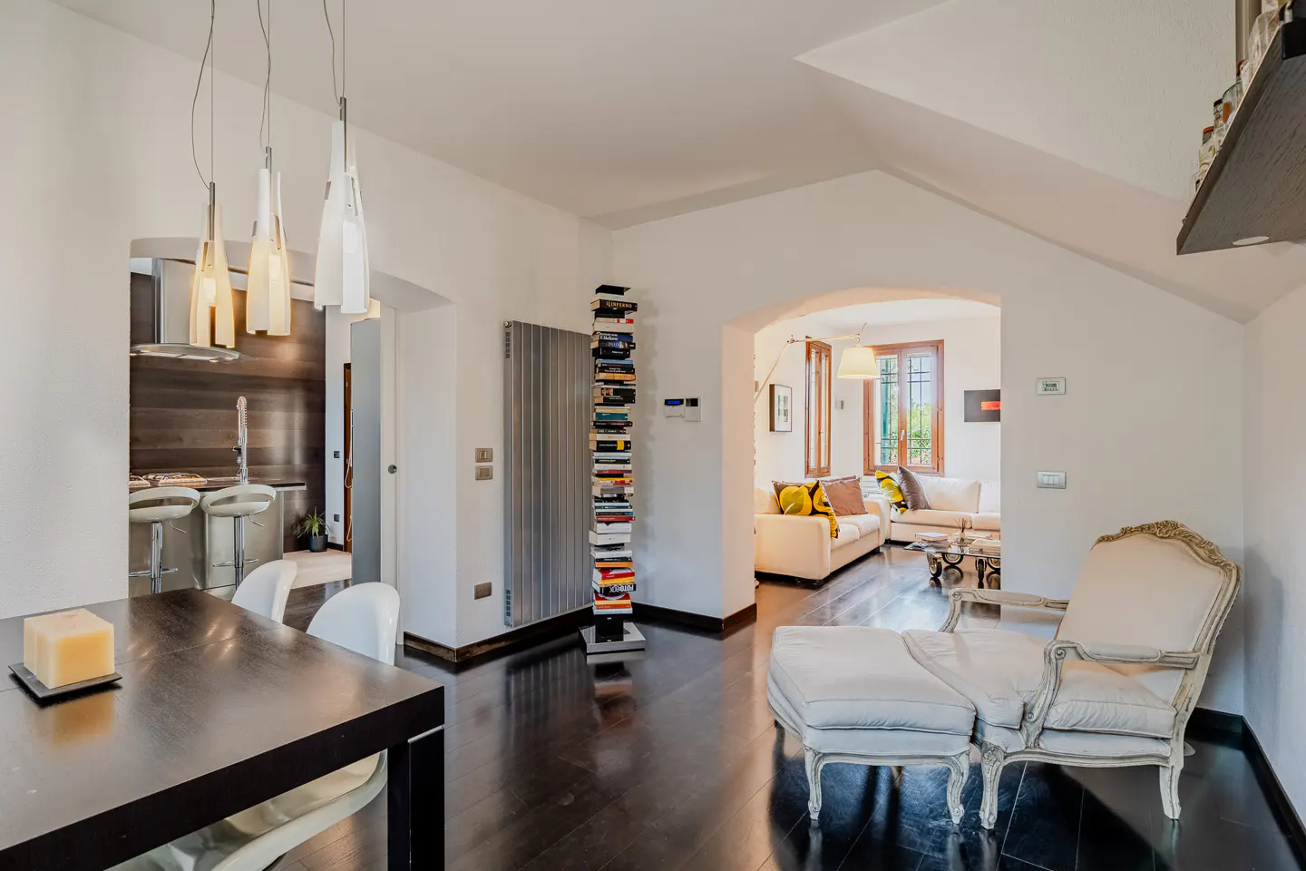 Interior view of a modern home with dark wood floors, white walls, and an open floor plan. A chaise lounge sits near an arched doorway.