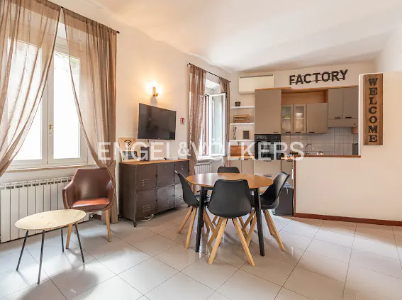 Open-plan living area with a dining table, black chairs, brown leather armchair, TV, and a modern kitchen with "FACTORY" sign.