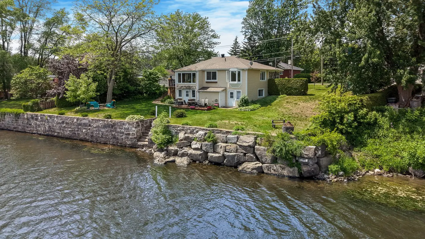 Two-story beige house with a grassy yard and stone retaining wall overlooking a river on a sunny day.