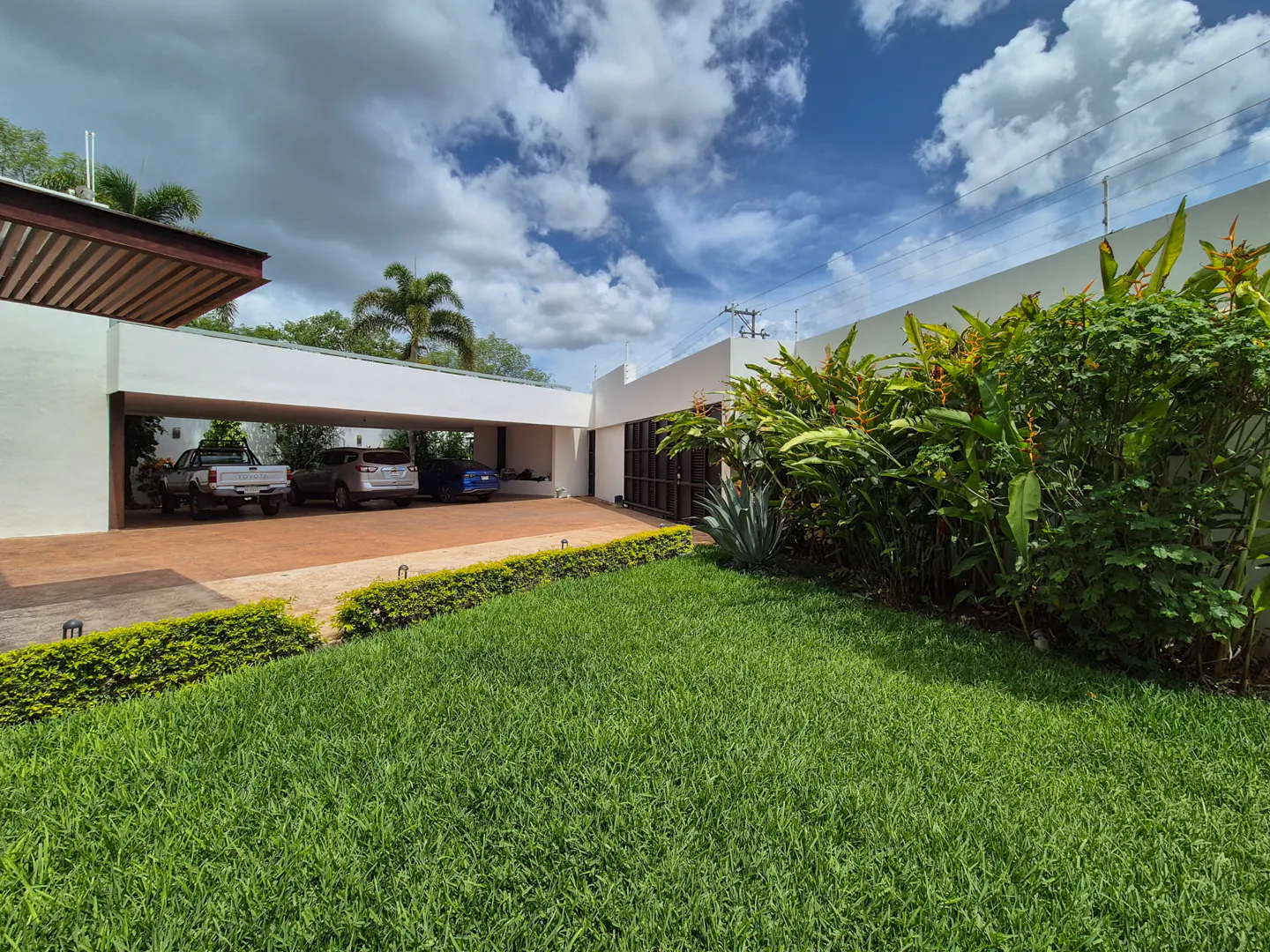 Modern white house with a carport holding three cars, a green lawn, and tropical plants under a cloudy blue sky.