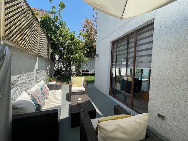 Outdoor patio with wicker furniture, striped cushions, and a small table. A sliding glass door leads inside. Green trees and a wooden fence are visible.