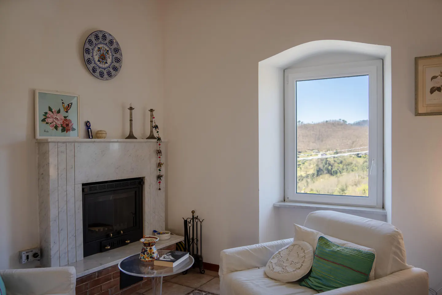 Living room with white walls, a marble fireplace, and a window with a view of green hills. A white armchair sits near a small table.