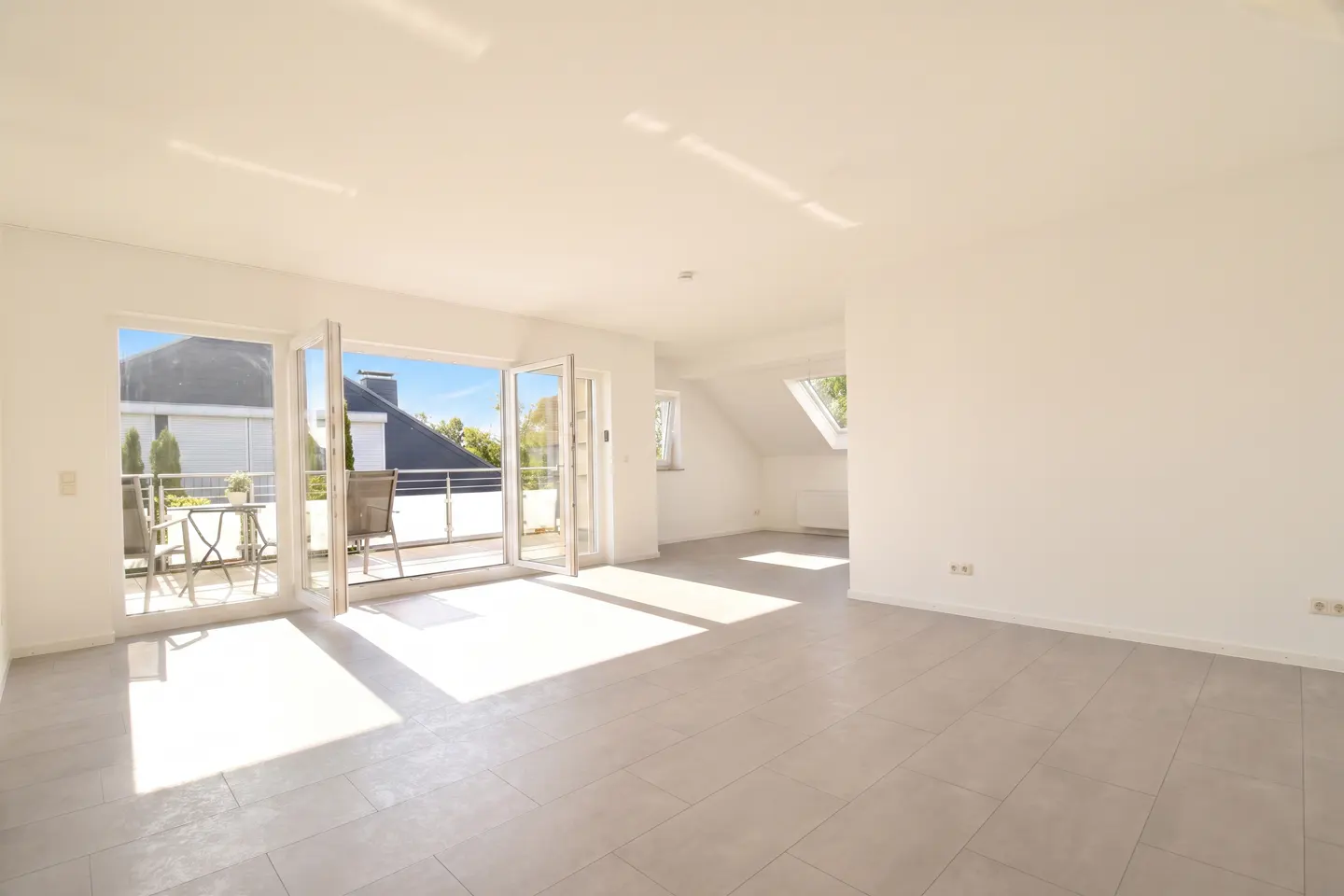 Bright, empty room with white walls and gray tile floor. Open glass doors lead to a balcony with outdoor furniture.