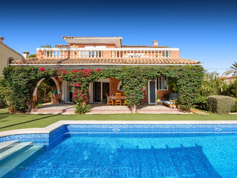 A terracotta house with a red tile roof is covered in green vines and red flowers, with a blue tiled pool in the foreground.