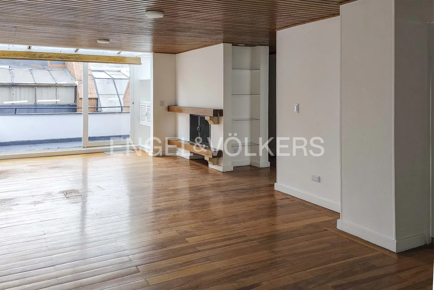 An empty living room with wood floors, a fireplace, and a large window leading to a balcony. The walls are white, and the ceiling is wood paneled.