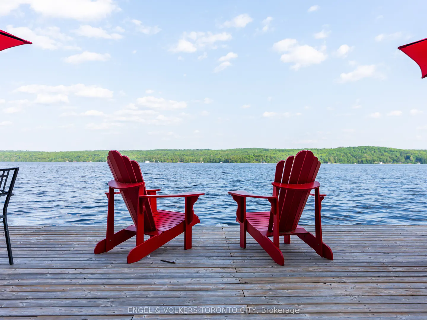 Two red Adirondack chairs face a lake on a wooden dock under a blue sky with white clouds.