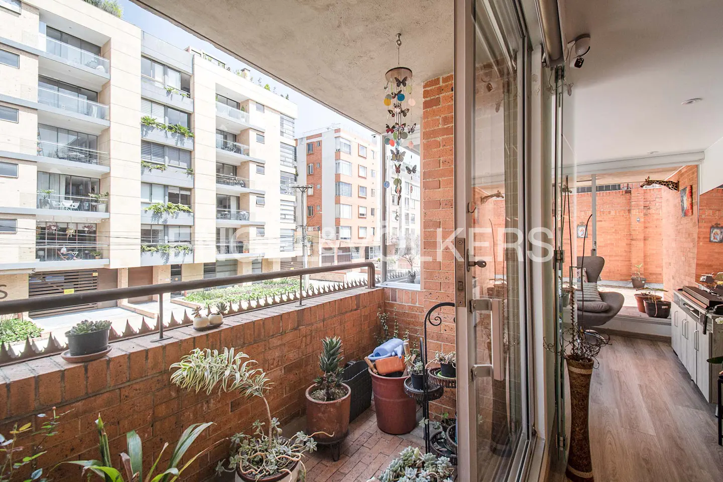 Balcony view with brick walls, potted plants, and a butterfly mobile. Apartment buildings visible in the background. Interior view through glass doors.