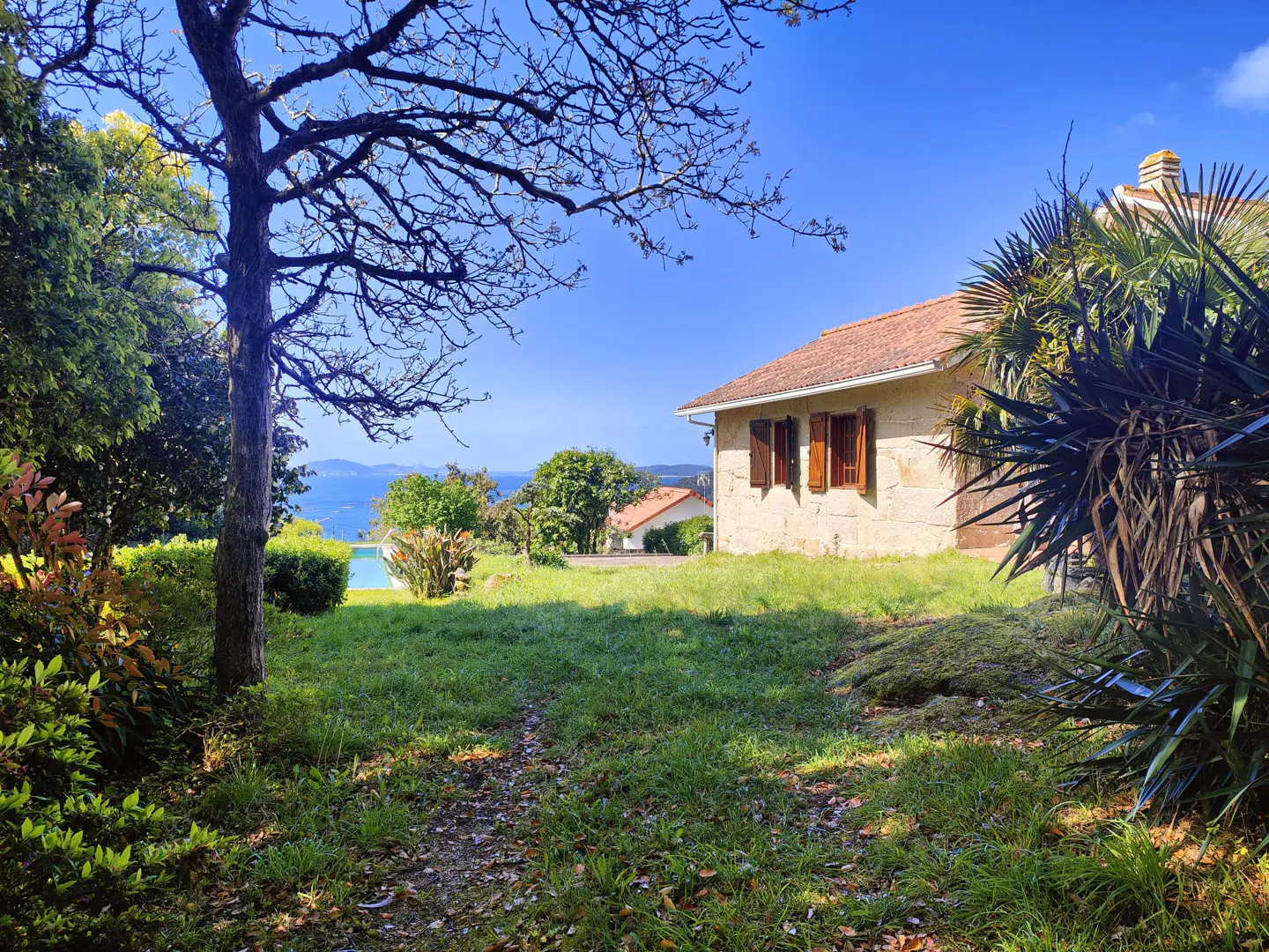 Exterior view of a stone house with a red tile roof, brown shutters, and a green lawn, with a tree and ocean in the background.