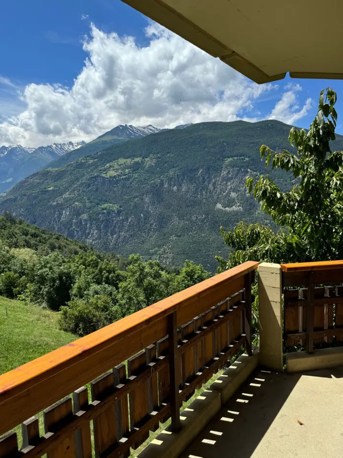 View from a balcony with a wooden railing overlooking green mountains and a blue sky with white clouds.