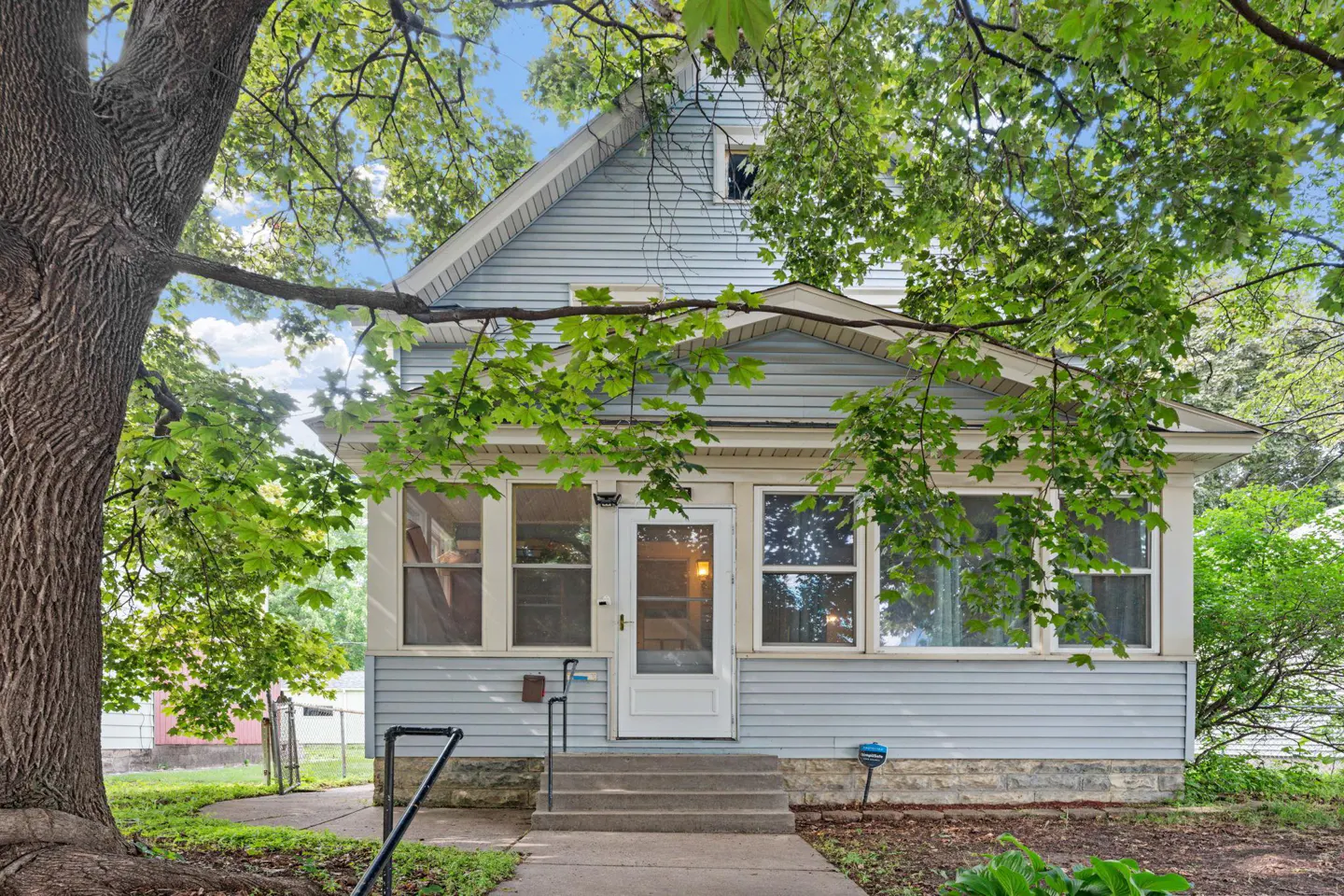 Light blue two-story house with a front porch, concrete steps, and large shade trees in the front yard.
