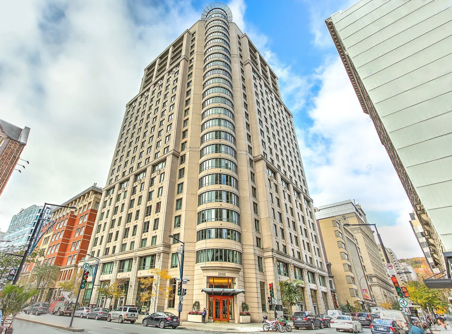 Exterior view of a tall, beige apartment building with many windows, a curved facade, and a glass-domed top, on a city street.