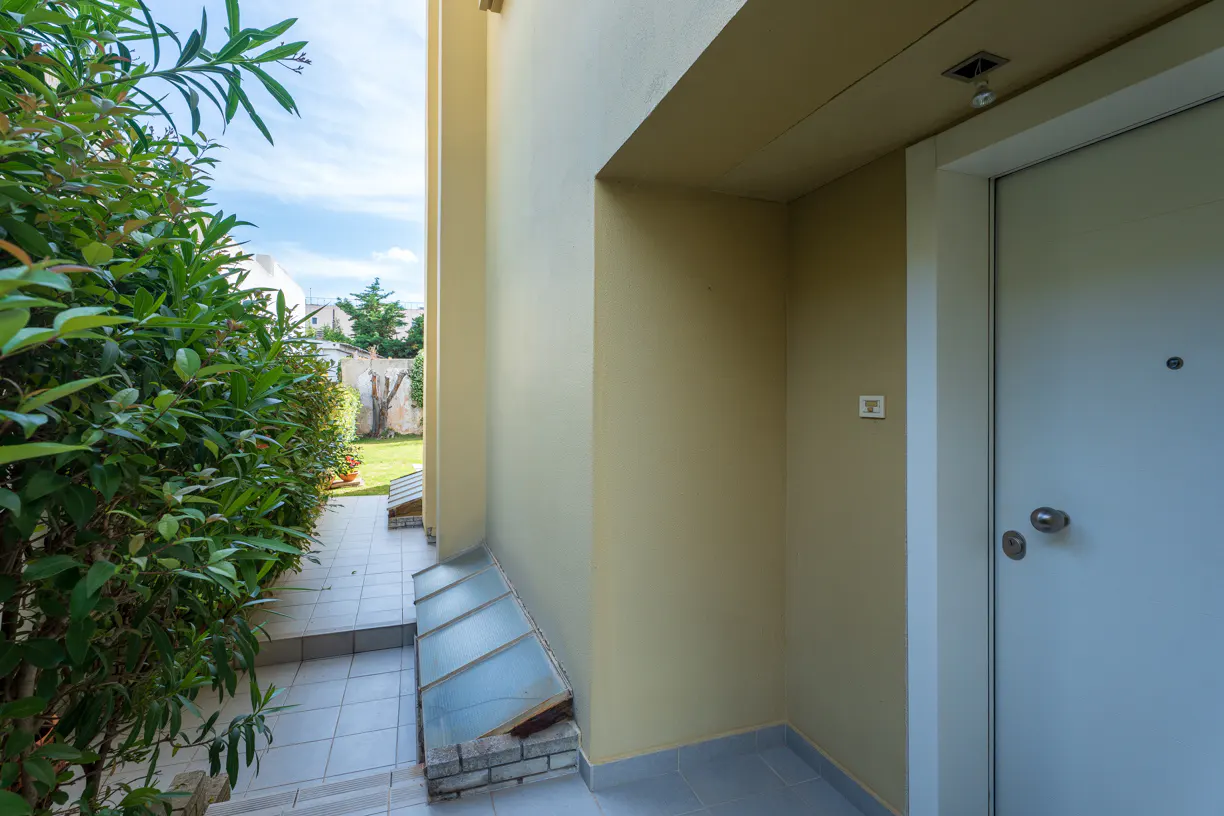 Exterior view of a home's entrance with a white door, beige walls, and green bushes along a tiled walkway.