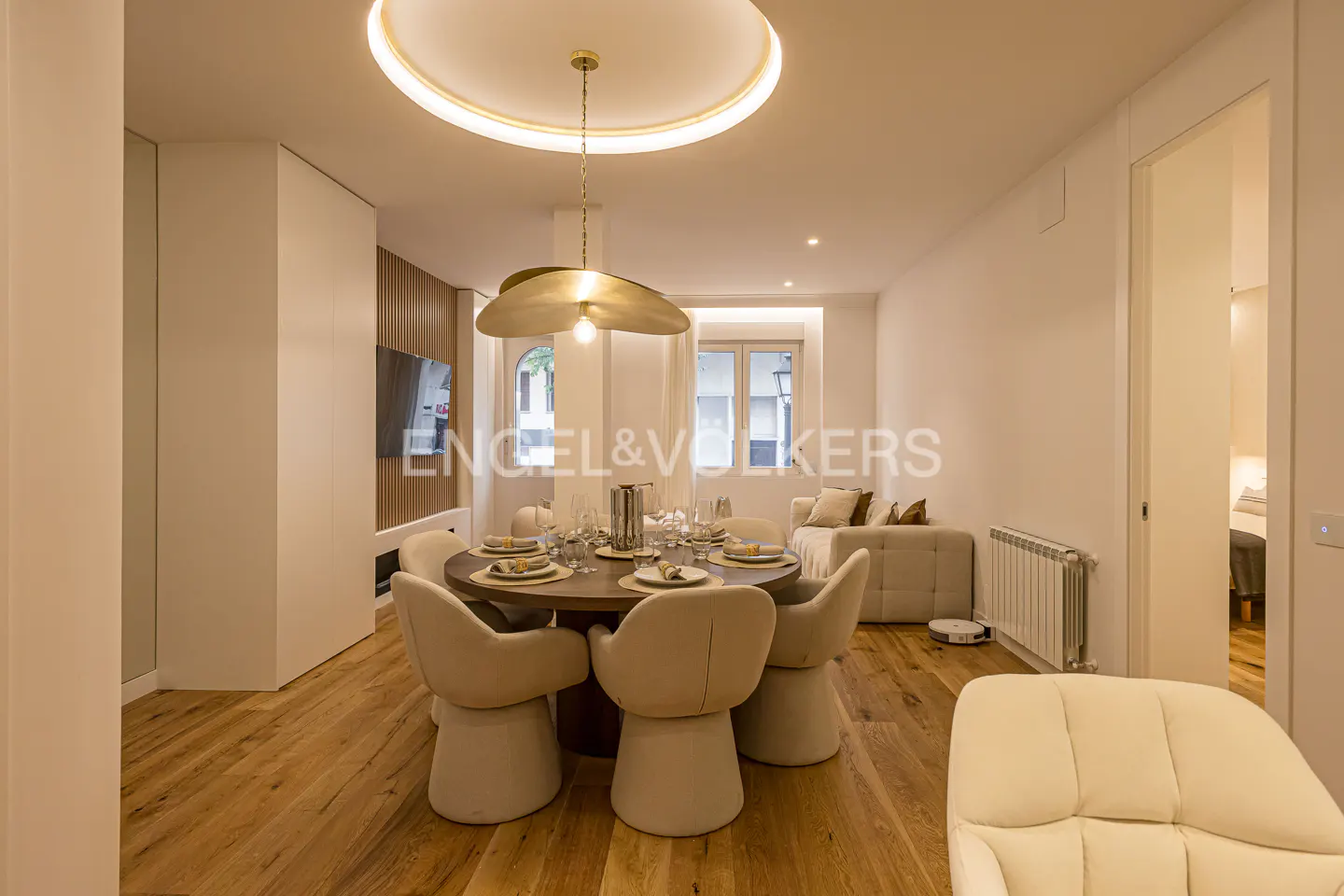 Modern dining room with a round wooden table, cream-colored chairs, a unique gold pendant light, and hardwood floors.